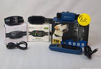 Three dog training collars displayed with their boxes and chargers: Dogrook humane no-shock bark collar, DINJOO TC03 Smart Bark Training Collar, Abbidot T30 E-Collar in blue.