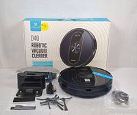 Photo of the DEENKEE D40 robotic vacuum cleaner with original box, charging dock, power adapter, side brushes, cleaning tool, and spare filters arranged in front.
