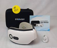 Eyeology Intelligent Eye Massager with carrying case, user manual, and charging cable displayed on a surface.
