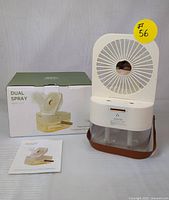 Photo showing the exact mini fan humidifier, its box, and user manual placed on a surface. The fan has a circular front with grill, transparent water reservoir, and control buttons.