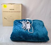 Photo showing the teal blue folded VEEMI heated blanket with attached white wired controller and original beige box with text and illustration.