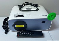 Front view of white XOPPOX mini projector with remote control and cables on top, showing lens capped and power plug cable coiled.