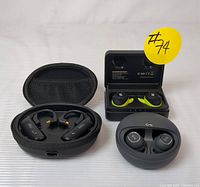 Image showing all three pairs of wireless earbuds with their charging cases. The Monster earbuds have a rectangular charging case and ear hooks. The TRN BT20S PRO earbuds are inside a zippered hard case. The third pair is in a rounded, gray charging case.