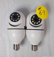 Two white CCTV cameras designed for E27 light socket with circular lens and multiple LED lights for night vision, shown side by side with a yellow sticker #69.