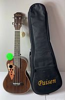 Front view of the Paisen Aquila ukulele next to its black carrying case with embroidered logo.