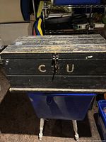 Front and top view of the black painted antique wooden storage box showing wear and chipping, with visible letter markings and metal latch.