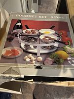 Closed original packaging box showing image of the cookware set with four pans cooking different foods, marketed for 4 persons.