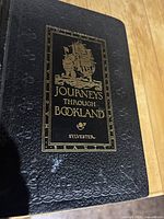 Close up of black textured cover with gold title Journeys Through Bookland and a sailing ship illustration.