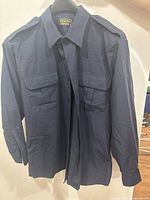 Navy blue long sleeved uniform shirt with two front flap pockets and shoulder epaulettes, hung on a black hanger.