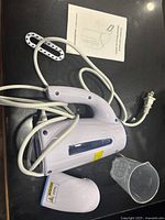White handheld electric garment steamer with cord wrapped, detachable nozzle attachment, and transparent measuring cup on black surface.