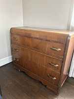 Front view of the brown wooden dresser with three drawers, showing carved accents and brass-tone metal handles. The finish has visible scratches and wear.