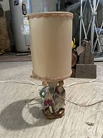 Full view of the ceramic table lamp with beige cloth shade and figurine base showing overall condition and design.