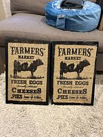 Two decorative wooden signs with burlap centers and black wooden frames showing farmers market theme graphics and text.