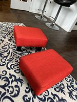 Pair of red fabric foot stools placed on patterned floor with visible plastic legs.