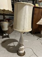 Full view of the lamp with white fabric cylindrical lampshade and off-white ceramic base with floral details on carved relief and gold accents.