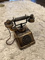 Vintage-style bronze-colored metal telephone replica standing upright on a soft, cream-colored textured surface, showing cradle, rotary dial, and cord attached to handset.
