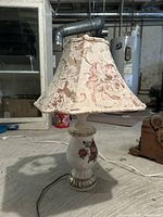 Full view of vintage ceramic table lamp on concrete floor, showing white base with red floral design and floral fabric lampshade.
