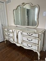 Full view of light beige wooden vintage vanity with scalloped details, drawers with brass handles, central cabinet doors, and matching large scalloped mirror above