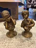 Two gold-toned vintage style bust sculptures, a girl and a boy, on a white carpeted floor with furniture in background