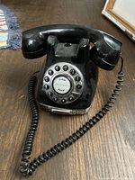 Front angle photo of black vintage rotary dial phone on wooden surface showing dial and coiled cord