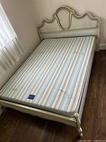 Full view of the full-size wooden bed frame in light beige paint with ornate curved headboard and footboard, mattress placed on frame, room with window and hardwood flooring.