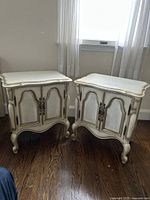 Two vintage wooden nightstands side by side in light beige color showing curved tops, scalloped edges, and ornate double doors with decorative handles.