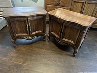 Pair of brown wooden nightstands showing front and side views, featuring curved tops, two paneled doors, brass decorative hardware, carved accents on bottom and curved legs.