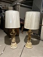 Pair of ceramic figurine table lamps with cream fabric lampshades seen from front in a room with dark furniture and concrete floor.