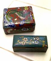 Top view of two cloisonné enamel boxes on white tile floor; larger red bird motif box alongside smaller green floral motif box.