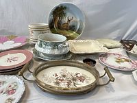 Overall view of various porcelain and pottery serve ware and decorative plates displayed on a white cloth, shows diversity and condition of items.