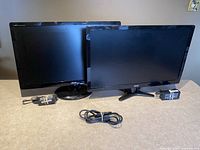 Two Acer widescreen computer monitors placed side by side on a table showing the black bezels, stands, power adapters, and one VGA cable in front.