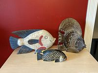 Three carved wooden fish sculptures displayed on a table against a red wall: large white fish with blue fins and red accent, medium dark fish with yellow accents, and tall abstract wooden fish sculpture.