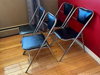 Four black vinyl and chrome folding chairs positioned against red wall and wooden floor, side view.