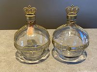 Front view of two clear glass Jacquin's Forbidden Fruit Liqueur bottles with gold-tone crown-shaped stoppers and decorative bands.