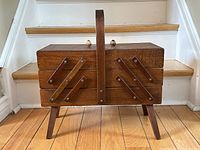 Closed vintage wooden accordion sewing basket showing dark wood finish, handle, angular legs, and brass hardware.