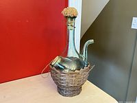 Front angled view of the vintage Italian glass decanter in wicker basket showing the long neck with cork, ice chamber inside, and woven wicker base.