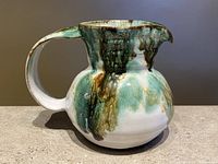 Front view of vintage ceramic mini creamer pitcher showing bulbous body and glazed dripping pattern in green and brown over white base.