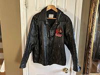 Front view of the black Redmen leather jacket on hanger. Shows Redmen patch on left chest, snap button front closure, collar, and two front angled pockets.