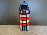 Front view of the Tiffany style stained glass lighthouse lamp illuminated, showing red, white, blue, and brown glass panels and black metal railings on the top.