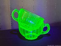 Two uranium glass teacups shown stacked, glowing bright green under UV light, highlighting the ribbed pattern and green handles.