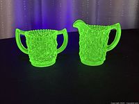 Two items, a sugar bowl and cream pitcher, glowing bright green under UV light against a dark background, showing intricate diamond pattern and handles.