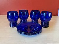 Four cobalt blue glass tumblers with rounded tops and ribbed bases, arranged behind four matching cobalt blue glass saucers on a light table against a red wall.