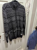 Full view of black and gray patterned wool sweater hanging, showing full zip front and ribbed collar and hem