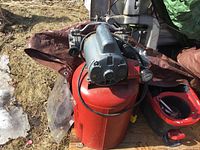 Air compressor viewed from the top-front showing the electric motor, compressor pump, attached pressure gauge and air hose. Compressor tank is painted red with signs of wear and rust. Located outdoors on a wooden pallet.
