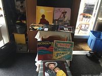 Five vinyl record albums displayed on a table and chair, showing colorful covers including Bonanza and Manhattan Transfer.