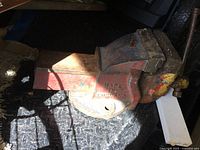 Heavy bench vise with worn paint and visible rust placed on a textured black surface, partially shaded by surrounding items.