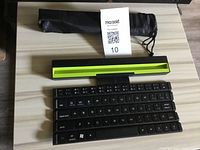 Black folding Bluetooth keyboard separated in three parts, next to a green and black device stand and a black carrying pouch on a light wood surface.