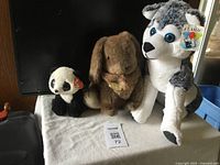 Three stuffed animals: brown plush rabbit with medallion, small black and white panda with Ty heart tag, and larger grey and white husky with Classic tag.