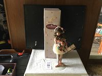 Full front view of the Ashton Drake doll standing with the box behind it on a white surface.