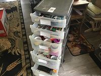 Gray rolling cart with partially open plastic drawers showing various craft supplies inside including ribbons, tapes, markers, and scissors.